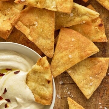 Air fryer pita chips on a wooden board along with a chip dunked in a bowl of hummus.