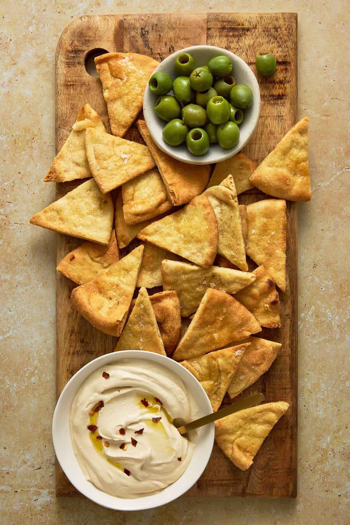 A wooden board filled with a pile of air fryer pita chips, a bowl of green olives and a bowl of plain hummus with a spoon in it.