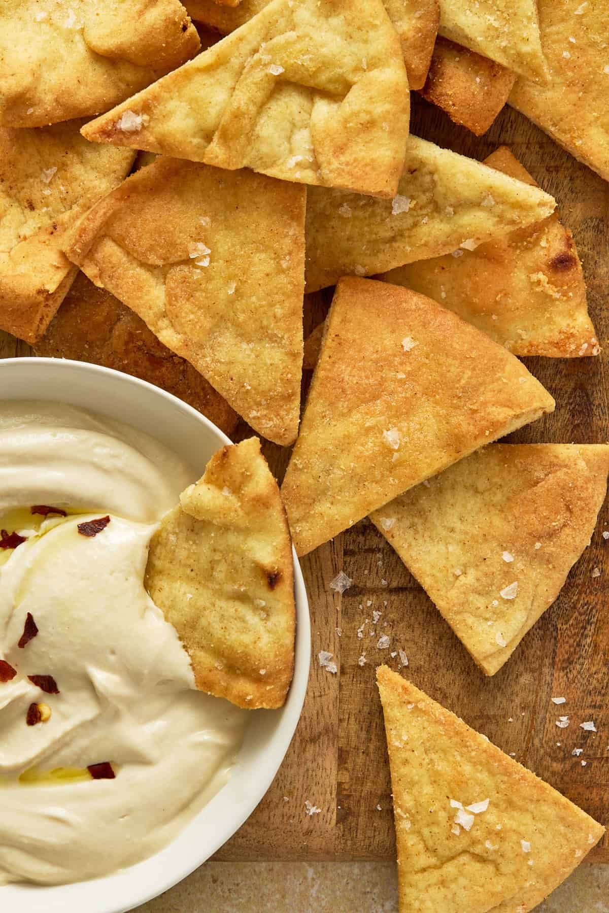 Air fryer pita chips along with a chip dunked in a bowl of hummus on a wooden board.