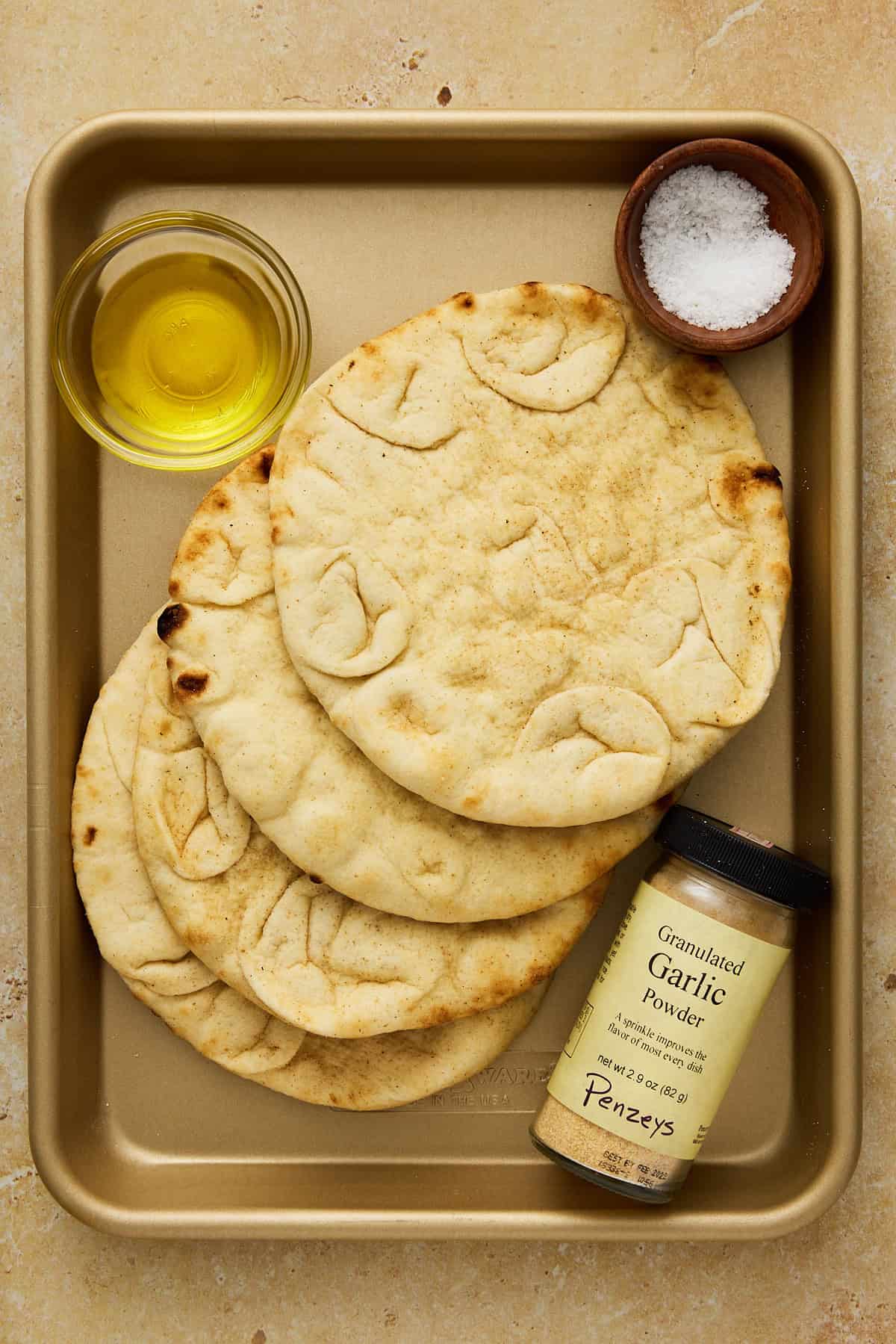 Fresh pitas, bowls of salt and olive oil and a container of garlic powder on a small gold sheet pan.