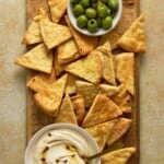 Wooden cutting board filled with green olives, air fried pita chips and a bowl of hummus.