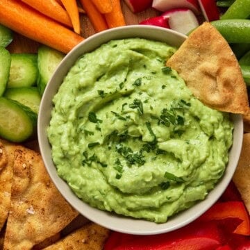 A light gray bowl filled with creamy avocado dip and sprinkled with fresh cilantro with a pita chip dunked in it.