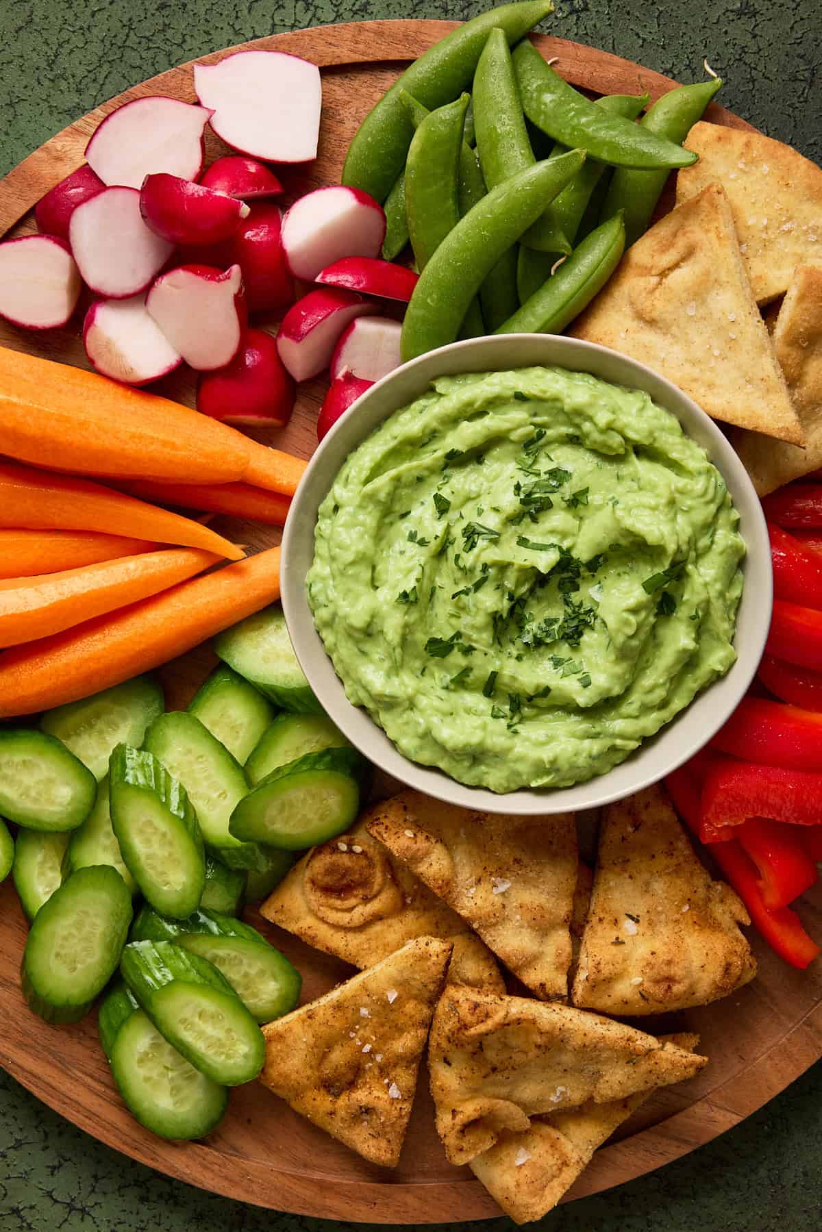 image of bowl of light green Avocado Cumin Yogurt Dip on wooden board surrounded by colorful vegetables and pita wedges. A round wooden cutting board filled with a fresh veggies, pita chips and a light gray bowl of creamy avocado dip.