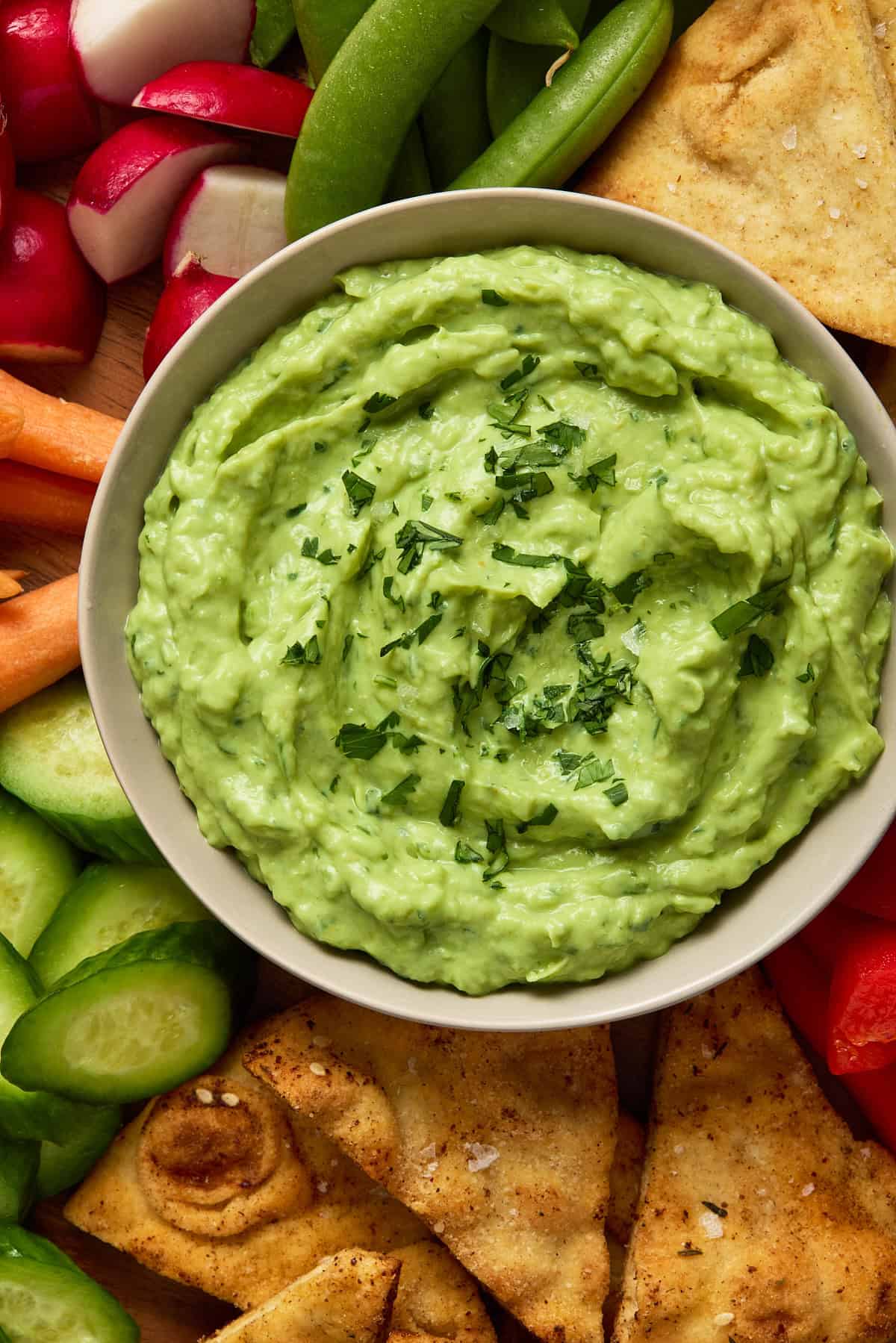 Creamy avocado dip in a light gray bowl on a wooden board with fresh veggies and pita chips.