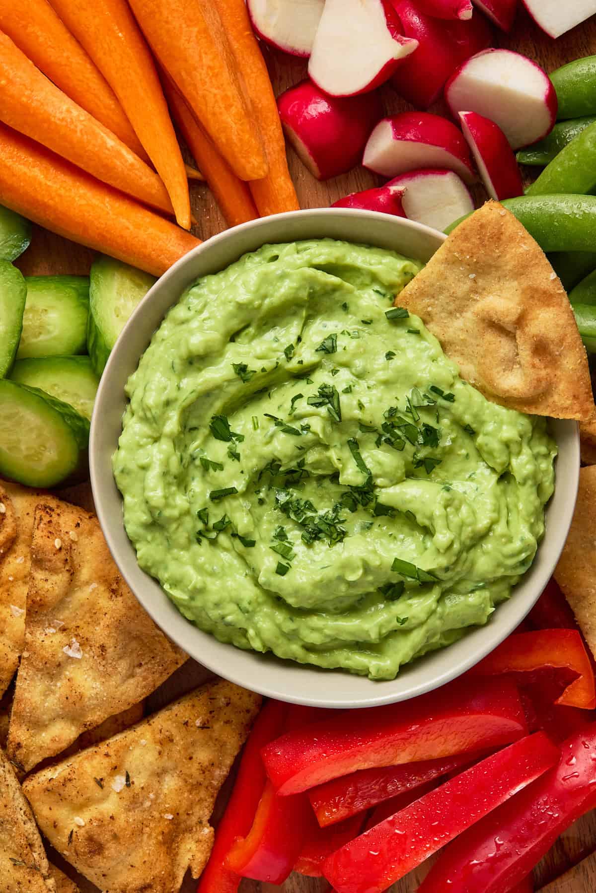 A pita chip dunked in a bowl of creamy avocado dip on a wooden board. Surrounded by fresh vegetables and more pita chips.