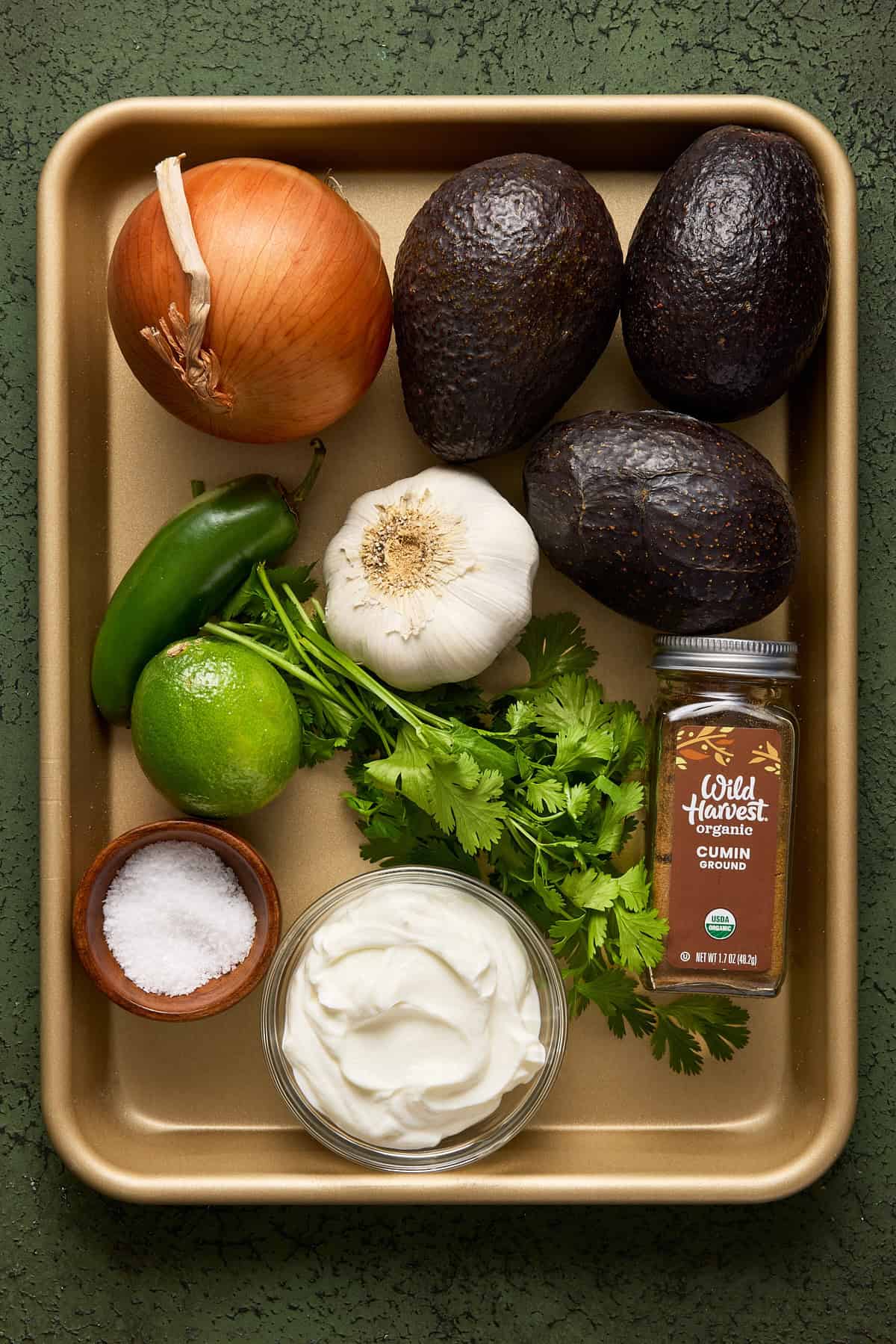 Ingredients for creamy avocado dip on a gold sheet pan including fresh avocados, onion, jalapeño, lime, cilantro, greek yogurt, cumin and salt.