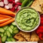 A bowl of creamy avocado dip on a wooden board surrounded by fresh veggies and pita chips.