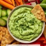 A pita chip stuck in a light gray bowl of creamy avocado dip surrounded by fresh veggies and pita chips.