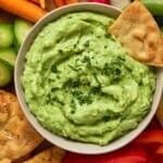 A bowl of creamy avocado dip with a pita chip dunked in it on a wood board with freshly cut veggies and more pita chips.