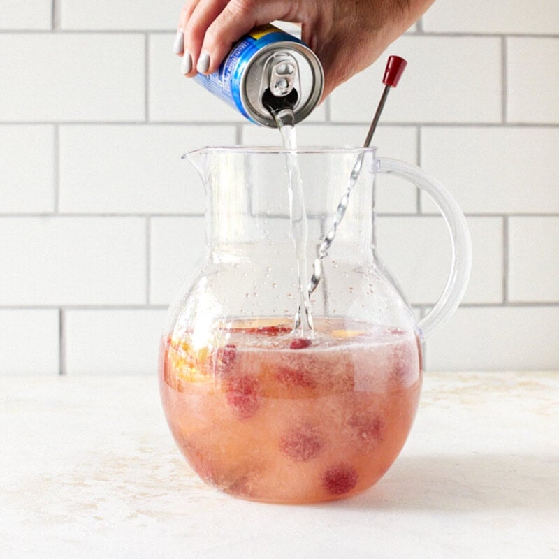 Club soda being poured into a glass pitcher filled with rosé sangria.