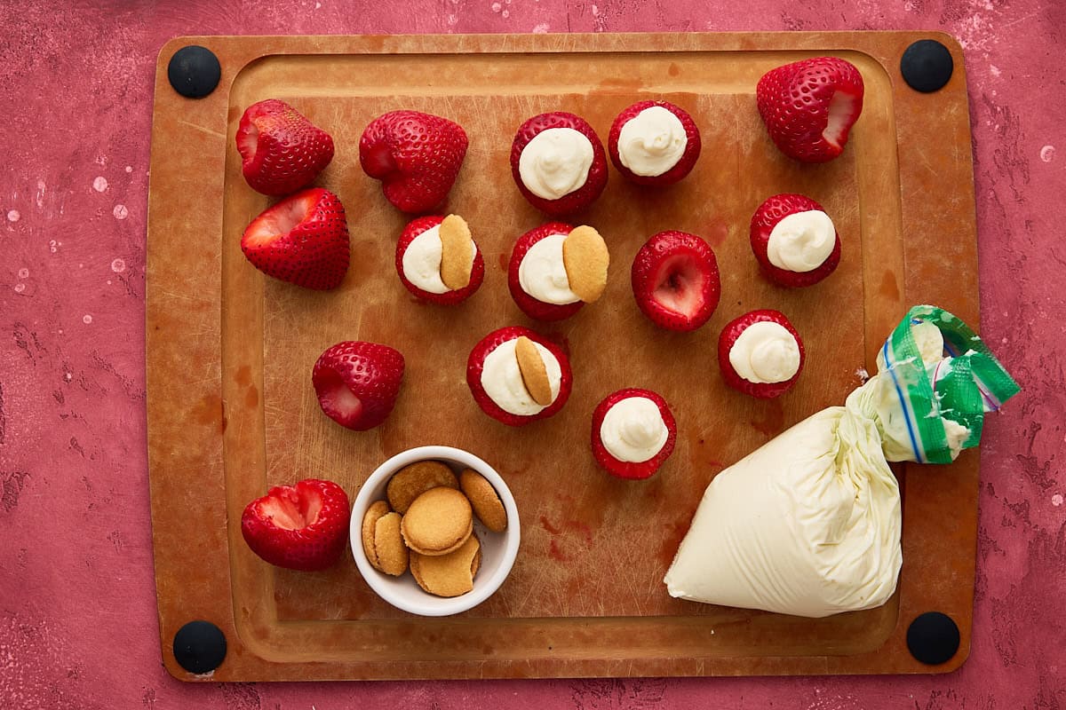 Strawberries filled with cheesecake filling on a cutting board with a bowl of mini Nilla wafers.