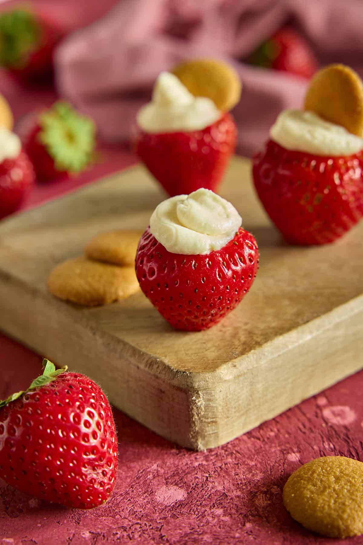 Cheesecake stuffed strawberries topped with mini Nilla wafers on a wood cutting board.
