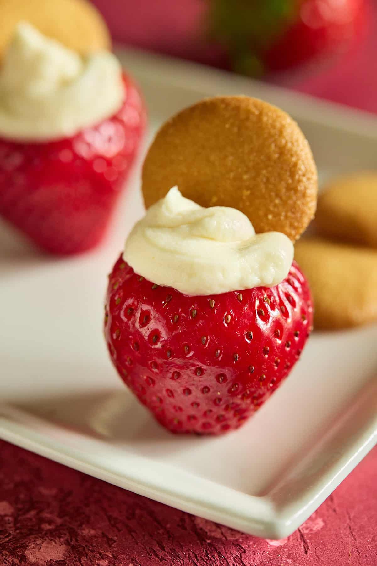 A hulled strawberry stuffed with cheesecake filling and topped with a mini Nilla wafer on a white plate.