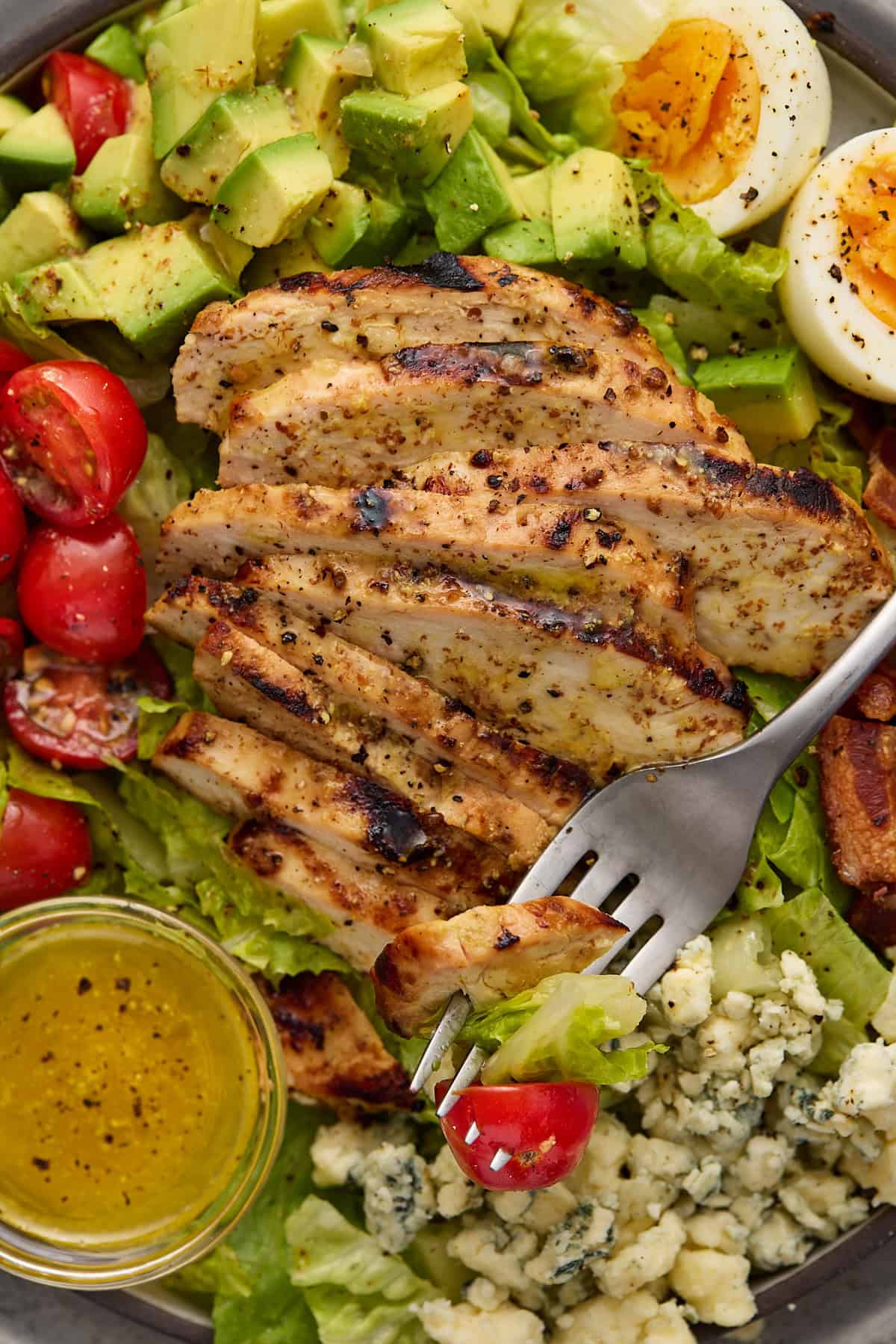 Close up imaged of sliced grilled chicken on top of cobb salad with a bite on a fork ready to eat.