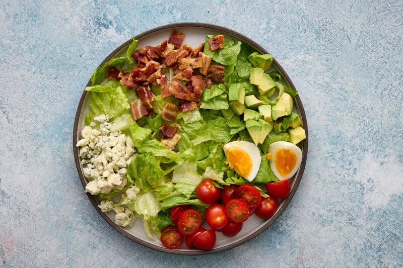 A plate filled with lettuce, bacon, avocado, hard boiled eggs, tomatoes and blue cheese.
