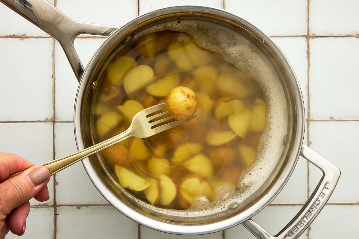 A hand holding a fork and spearing a par boiled potato half in a pot of water.