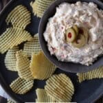 Green olive dip in a bowl on a gray plate with potato chips.