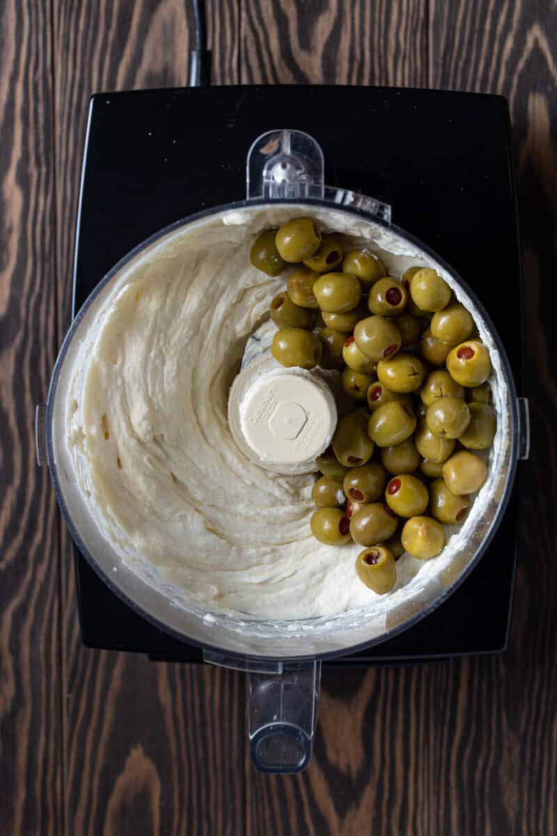 Green olives on top of smooth cream cheese mixture in food processor.