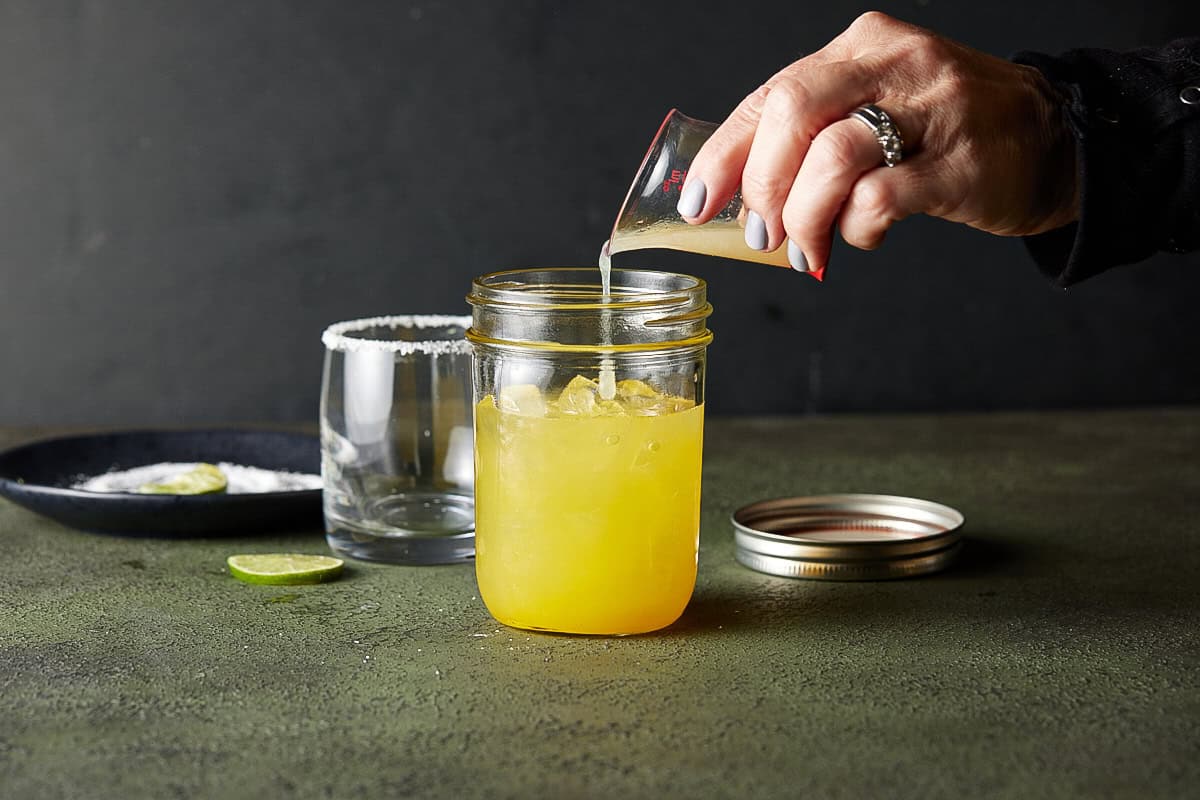 Hand pouring juice into a mason jar filled with mango margarita.