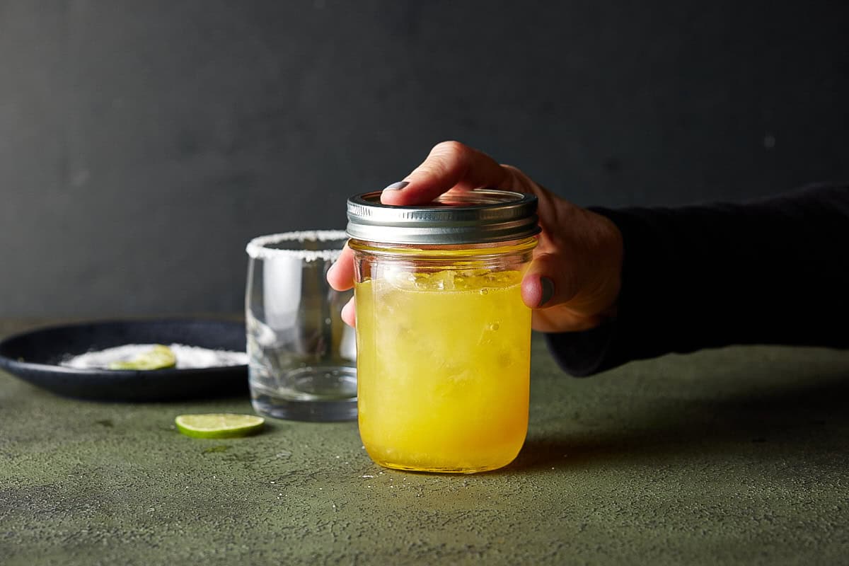 Hand on a closed mason jar filled with ice and mango margarita.