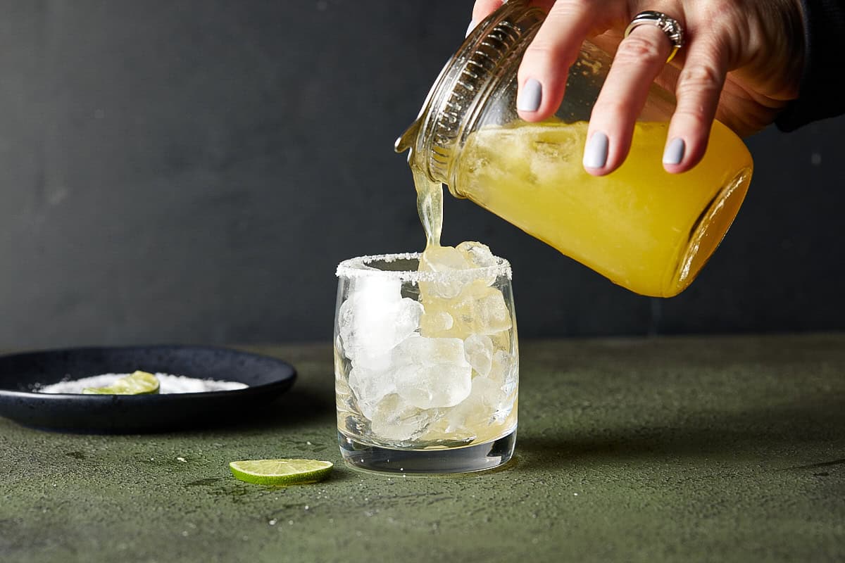 Hand pouring mango margarita into a prepared lowball glass from a mason jar.