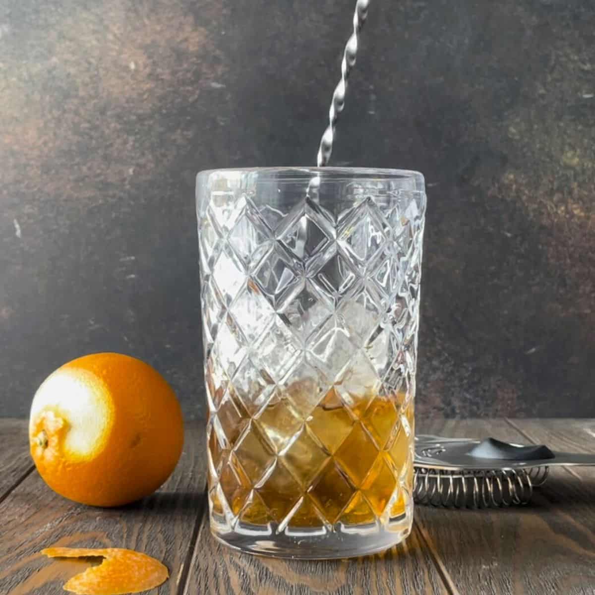 Hand holding a bar spoon and stirring añejo tequila old fashioned cocktail in a mixing glass.
