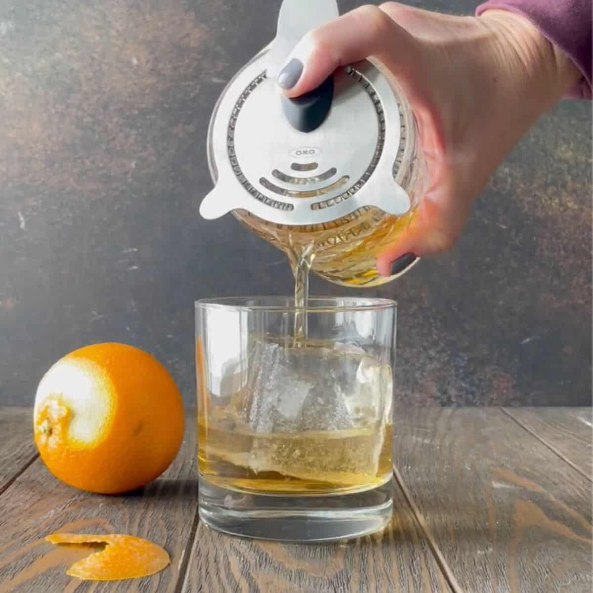 Hand straining añejo tequila old fashioned cocktail into a lowball glass with a large ice cube.