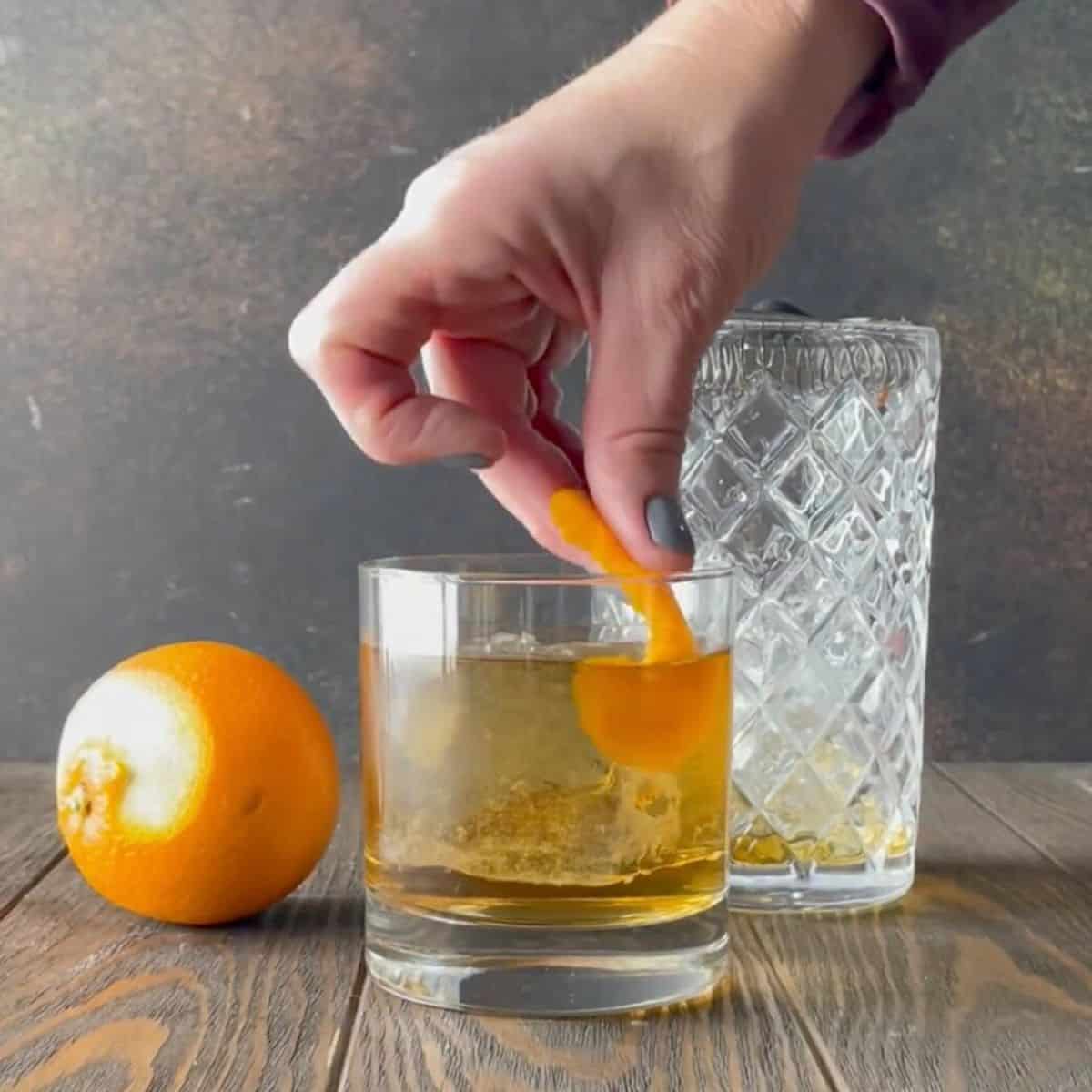 Hand dropping an orange peel into a lowball glass filled with añejo tequila old fashioned cocktail.