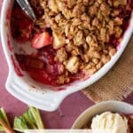 A spoon in a white baking dish filled with apple rhubarb crisp.