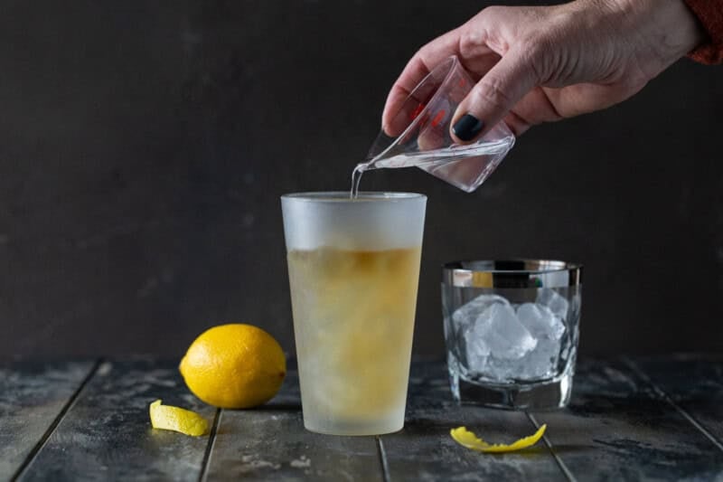 Hand pouring simple syrup into a glass cocktail shaker filled with bourbon, elderflower liqueur, lemon juice and ice.