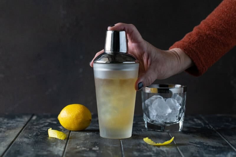 Hand on a glass cocktail shaker filled with bourbon elderflower cocktail next to an ice-filled lowball glass.