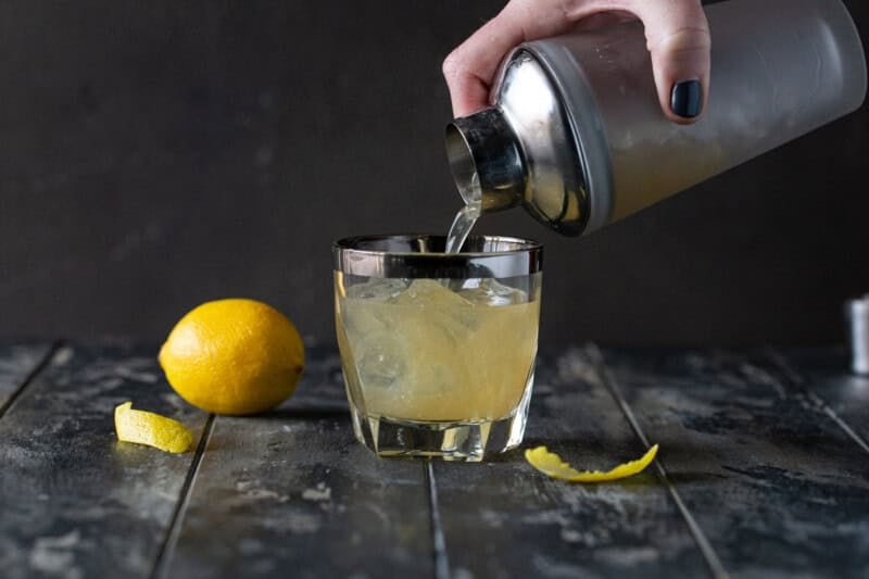 Hand holding a cocktail shaker and pouring a bourbon elderflower cocktail into a lowball glass filled with ice.