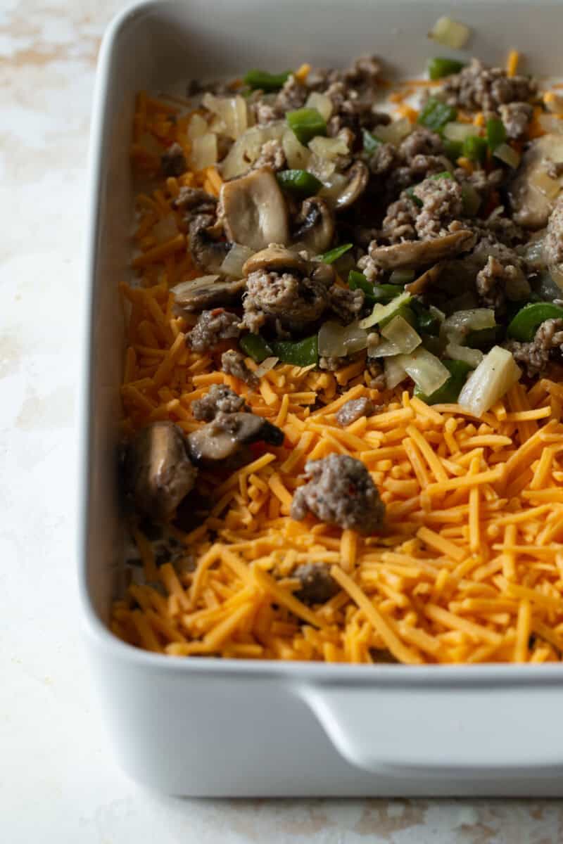 Layers of shredded cheddar cheese, cooked sausage, mushrooms and green bell pepper in a white casserole dish.