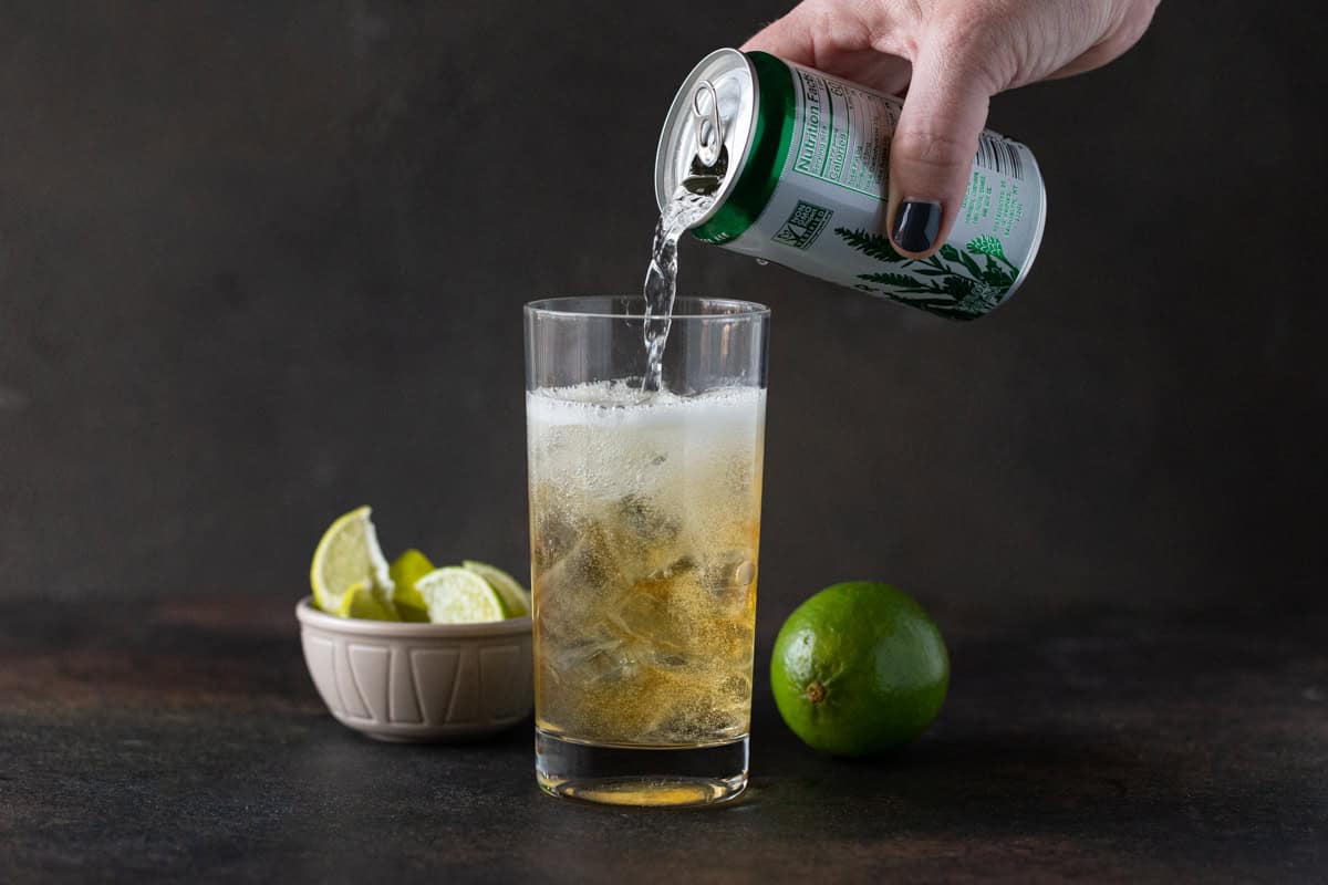 Hand pouring ginger ale into a highball glass filled with ice and whiskey.