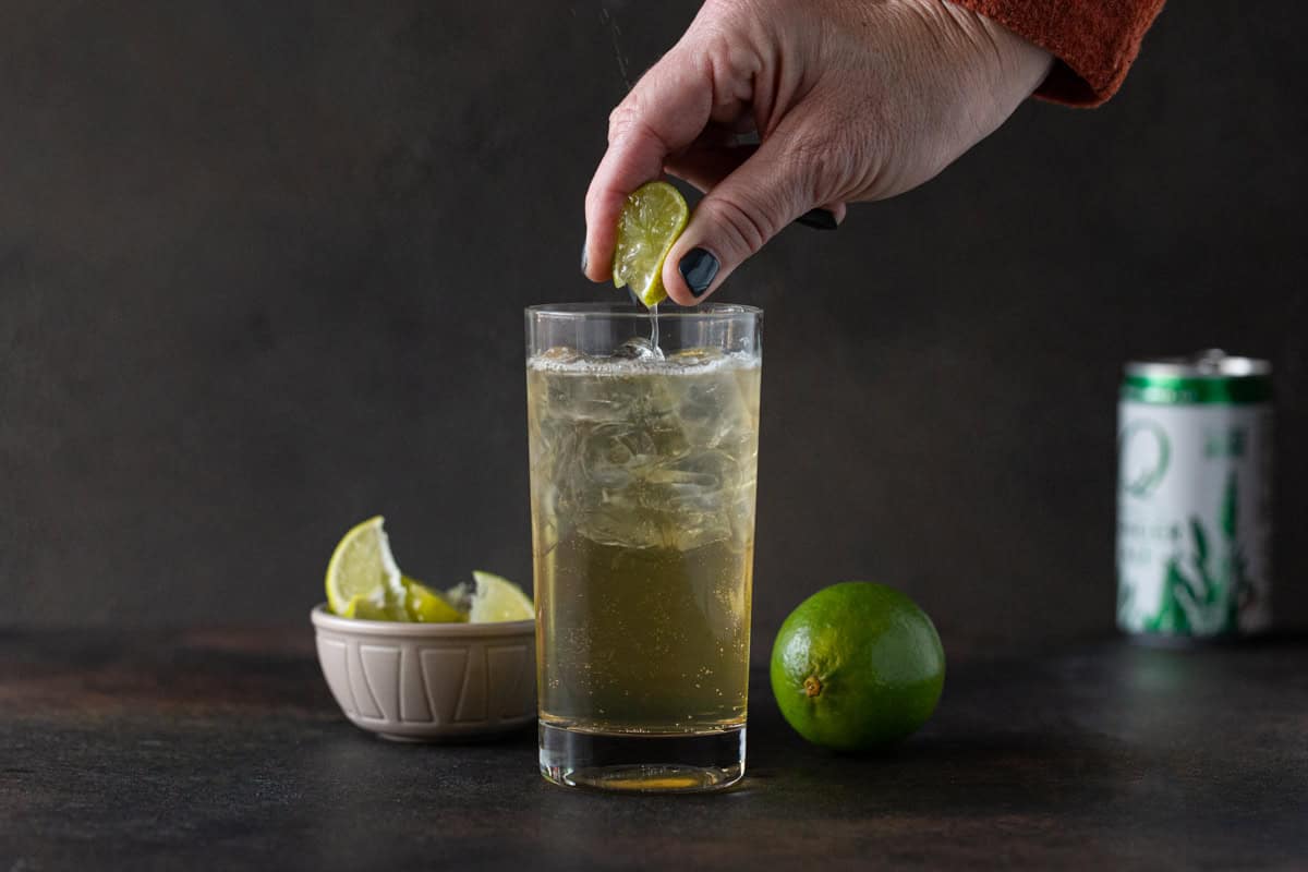 Hand squeezing a lime wedge into a highball glass filled with ginger whiskey cocktail.