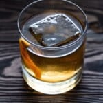 Lowball glass of añejo tequila old fashioned with a single square ice cube and garnished with an orange peel.