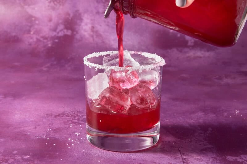 Shaker straining blueberry margarita into salted glass.