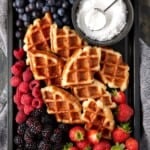 Croissant waffles piled on a dark sheet pan and surrounded by fresh berries and a bowl of powdered sugar.