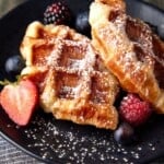 Two croissant waffles and berries on a black plate dusted with powdered sugar.