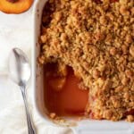 peach crumble in a white pan with a spoon and fresh peaches.