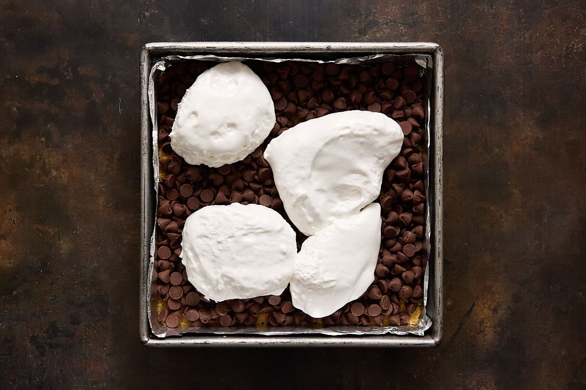 Dollops of marshmallow creme on top of chocolate chips and a graham crust in a square metal pan.