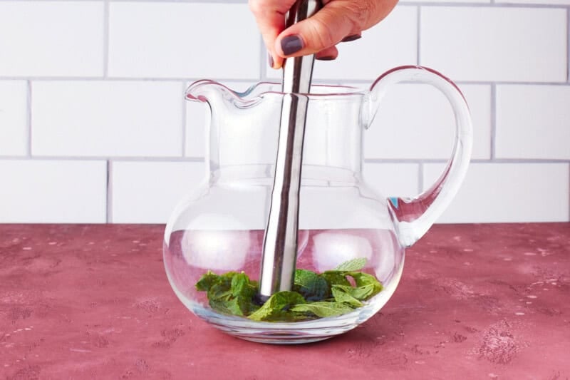 Hand muddling mint leaves and lime juice in a glass pitcher.