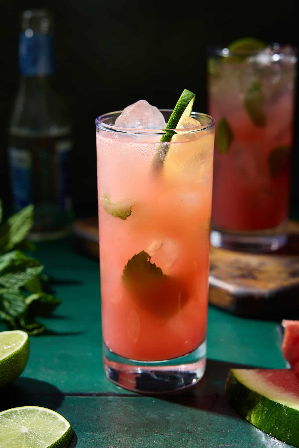A watermelon mojito cocktail in a highball glass. Surrounded by fresh mint, lime and watermelon wedges.