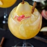 A round wine glass filled with apple cider sangria and garnished with slices of apple, pear and cinnamon sticks.
