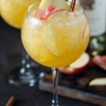 Round wine glass filled with apple cider sangria, apple slices, a pear slice and cinnamon stick.