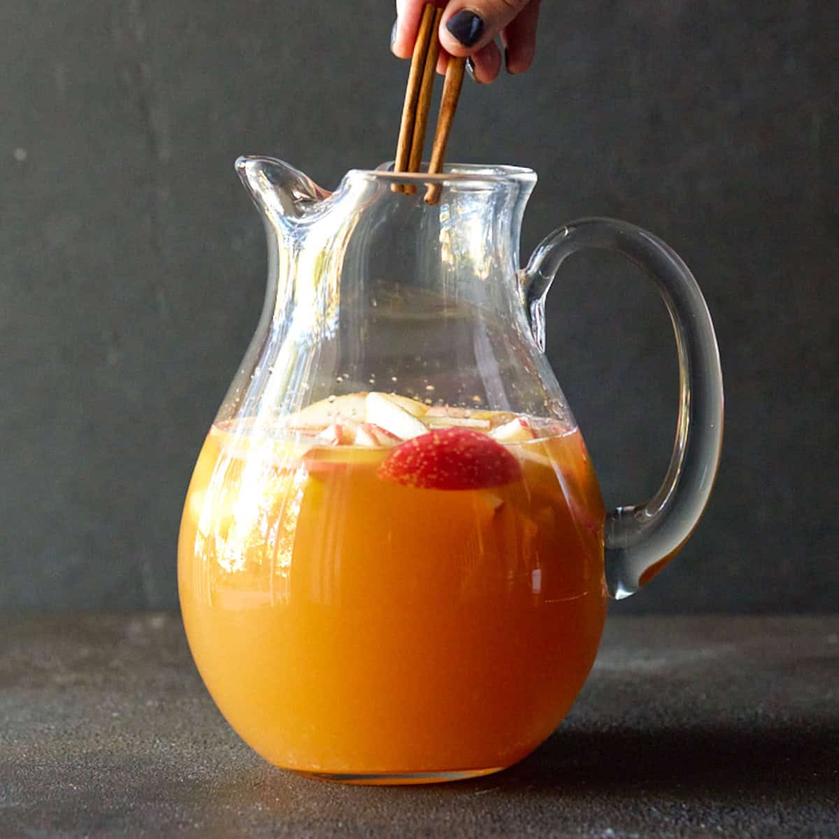 Cinnamon sticks being added to a glass pitcher of apple cider sangria.