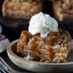 An apple crisp bar on a wooden plate topped with a scoop of vanilla ice cream and drizzled with caramel sauce.