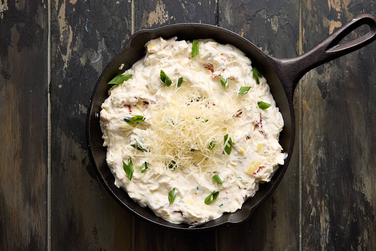 Artichoke dip in cast iron pan ready to bake with shredded parm on top.