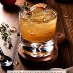 A glass of bourbon apple cider cocktail on a wooden coaster. Garnished with thin apple slices and a sprig of fresh thyme.