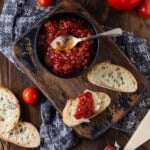 A bowl of homemade tomato jam surrounded by baguette slices that are topped with parmesan cheese and a dollop of tomato jam.